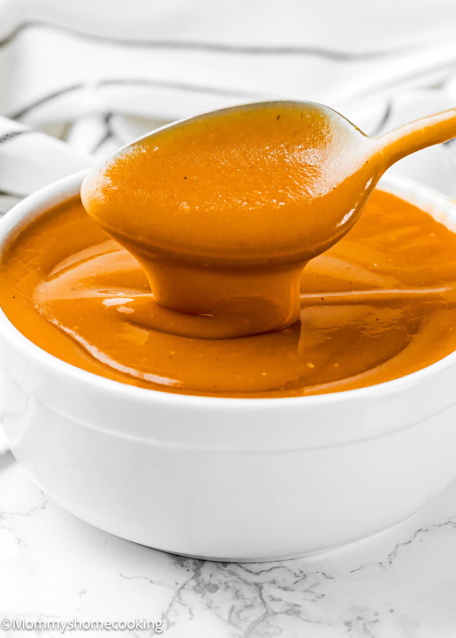 A white bowl filled with smooth, orange, no dripping flavorful gravy, with a spoon lifting some of the sauce above the bowl.