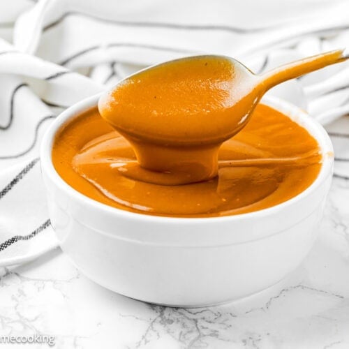 A spoon holds up thick, orange-brown flavorful gravy above a white bowl filled with the same no dripping gravy, set on a marble surface with a striped cloth in the background.