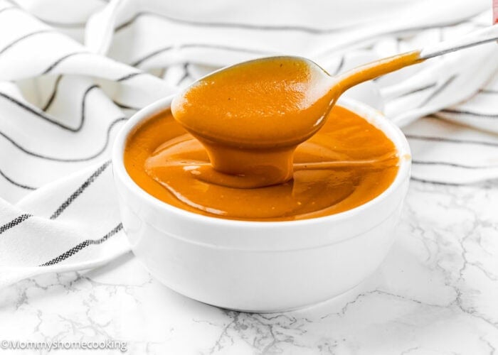 A spoon holds up thick, orange-brown flavorful gravy above a white bowl filled with the same no dripping gravy, set on a marble surface with a striped cloth in the background.