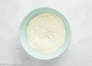 A light blue bowl filled with a mound of white powdered flour sits on a white textured surface, ready for baking Cranberry Orange Muffins without Eggs.