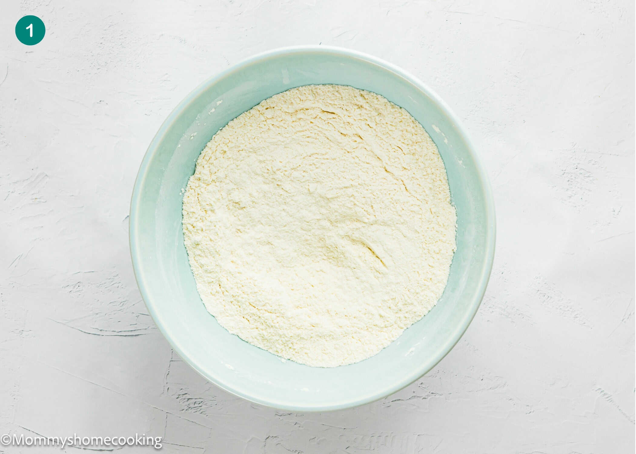 A light blue bowl filled with flour sits on a white textured surface, ready for making Cranberry Orange Muffins without Eggs.