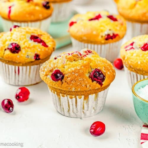 A group of Cranberry Orange Muffins without Eggs on a white surface, with scattered cranberries and bowls of sugar nearby.