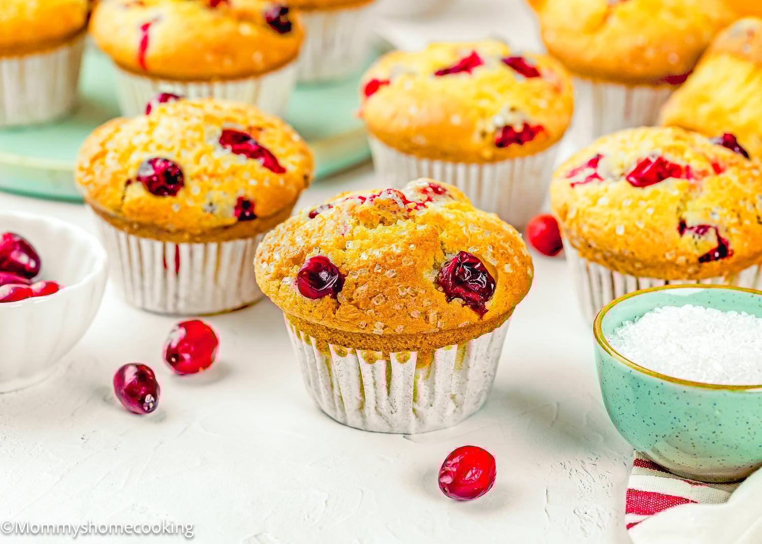 A group of Cranberry Orange Muffins without Eggs on a white surface, with scattered cranberries and bowls of sugar nearby.