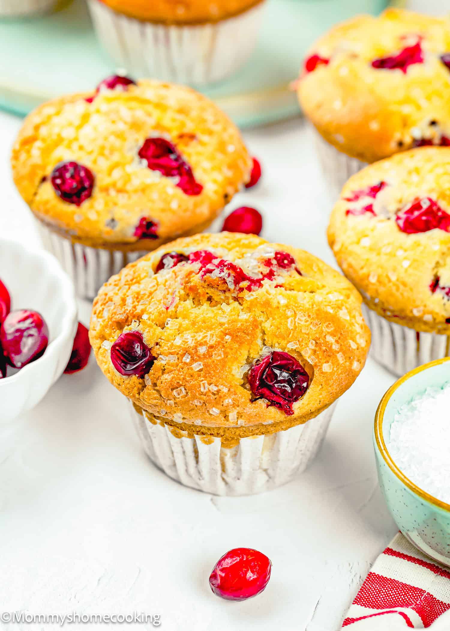 A close-up of freshly baked Cranberry Orange Muffins without Eggs in paper liners, with whole cranberries and a bowl of sugar nearby on a white surface.