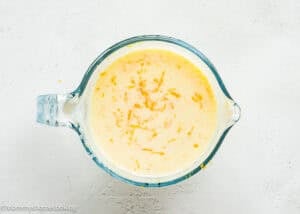 A glass measuring cup filled with a creamy yellow mixture for Cranberry Orange Muffins without Eggs, topped with orange zest, on a light textured surface.