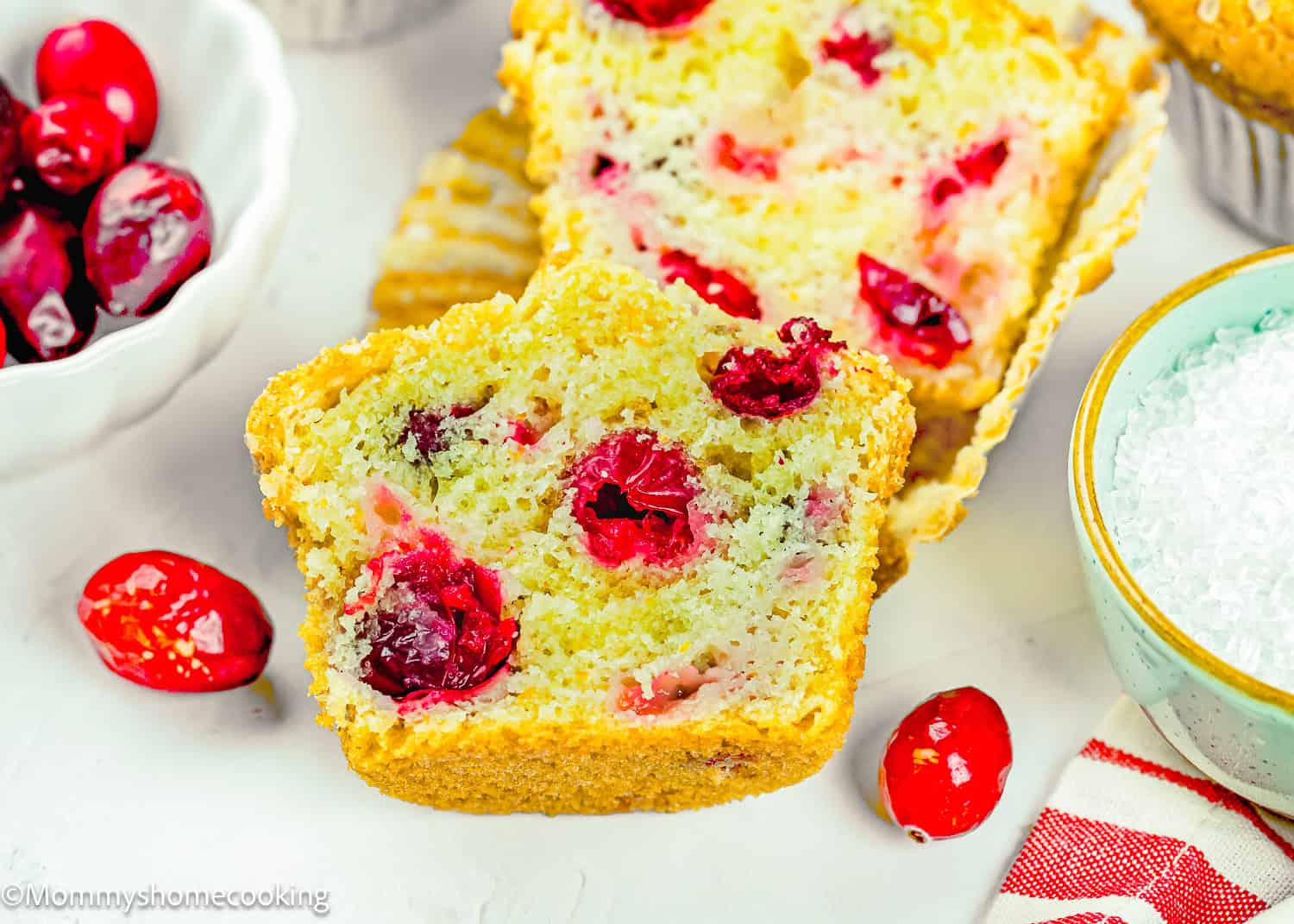 A close-up of sliced Cranberry Orange Muffins without Eggs with visible cranberry pieces, next to whole cranberries in a bowl and a white bowl of sugar on a white surface.
