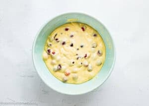 A light blue mixing bowl filled with thick, yellow batter for Cranberry Orange Muffins without Eggs, containing whole grapes and cranberries, placed on a white textured surface.