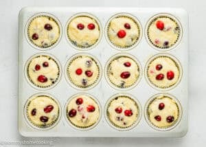 Muffin tin filled with 12 unbaked Cranberry Orange Muffins without Eggs, each portion topped with cranberries, on a white background.