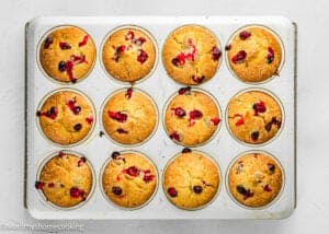 A muffin tin containing twelve freshly baked Cranberry Orange Muffins without Eggs on a white surface.
