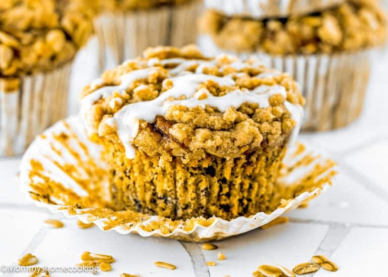 A close-up of Eggless Banana Crumb Muffins, streusel-topped with an icing drizzle, partially unwrapped in a white paper liner and surrounded by other muffins.