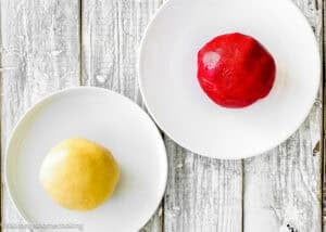 Two round balls of dough, one yellow and one red, sit separately on white plates atop a rustic white wooden surface, ready for making Eggless Candy Canes Cookies.