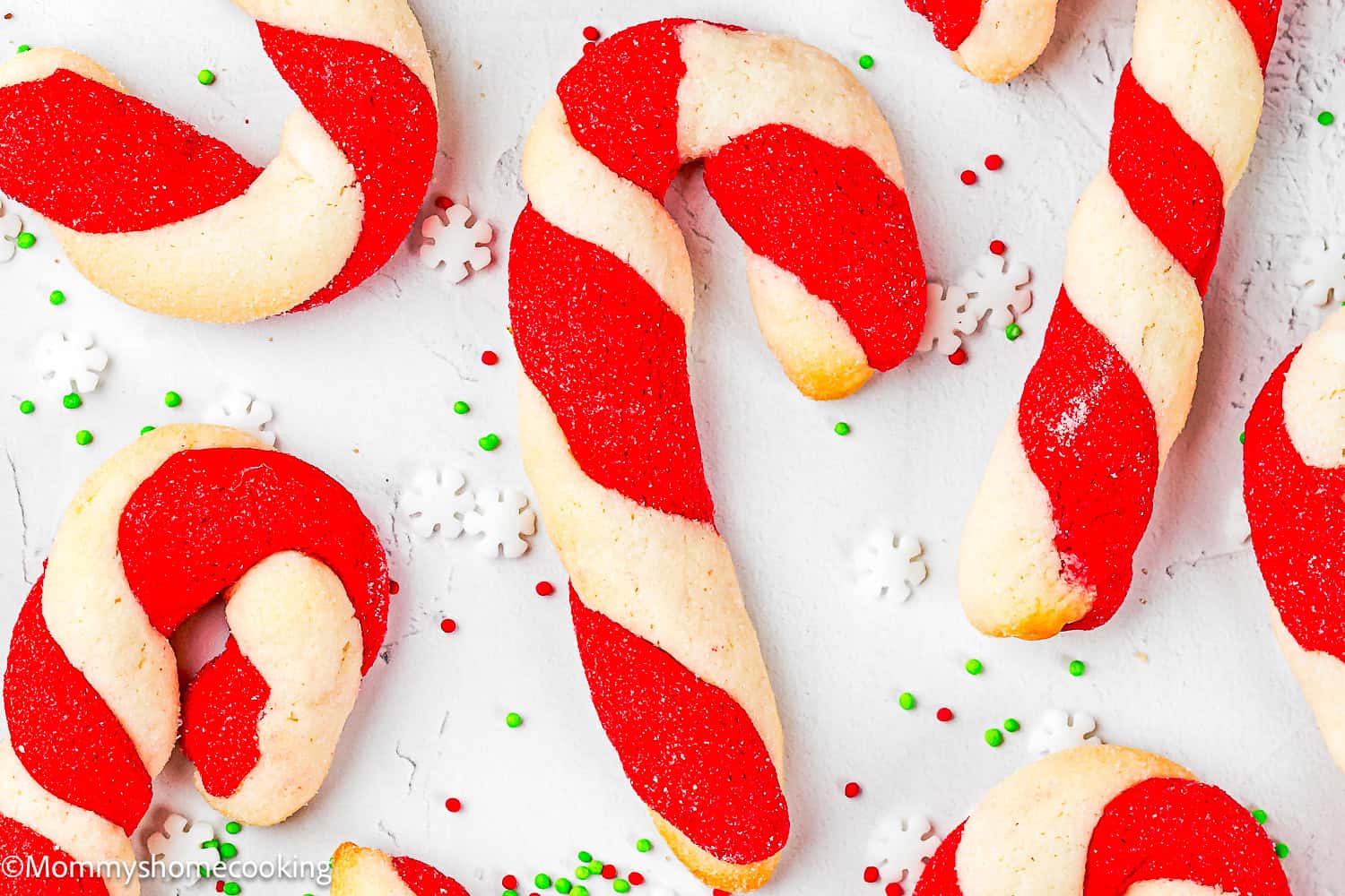 Eggless Candy Canes Cookies with red and white twisted dough are arranged on a white surface, surrounded by small green, red, and white sprinkles.