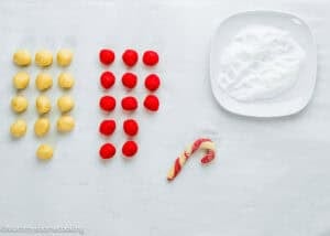 Rows of yellow and red dough balls, a plate of sugar, and a partially shaped Eggless Candy Canes Cookies on a white surface.