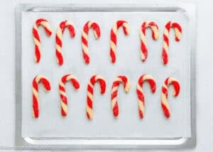 A baking sheet with parchment paper holds twelve Eggless Candy Canes Cookies, shaped in red and white candy cane stripes and arranged in neat rows.