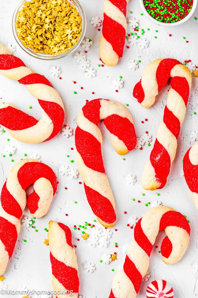 Eggless Candy Canes Cookies with red and white sugar stripes are arranged on a white surface, surrounded by small bowls of sprinkles and festive decorations.