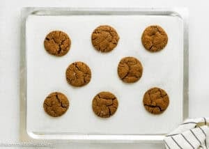 Nine round, cracked Eggless Chocolate Ginger Cookies are arranged in rows on a parchment-lined baking sheet, viewed from above. A striped towel is visible in the bottom right corner.