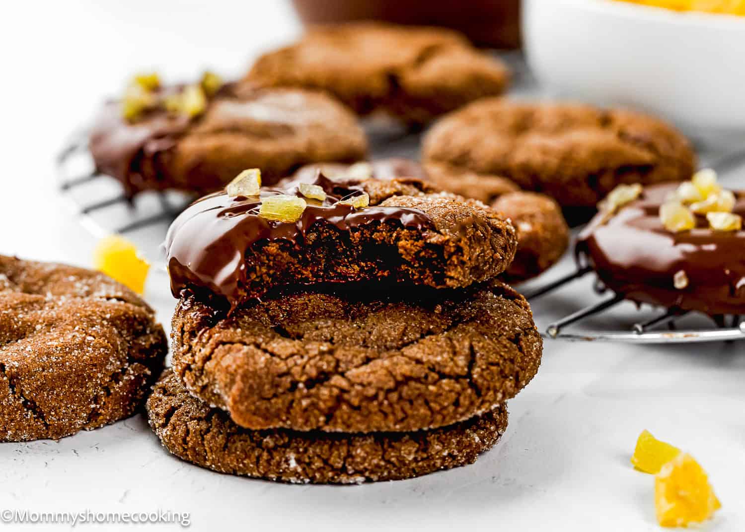 A stack of Eggless Chocolate Ginger Cookies, with one partially dipped in chocolate and topped with candied ginger, sits next to more cookies on a cooling rack.