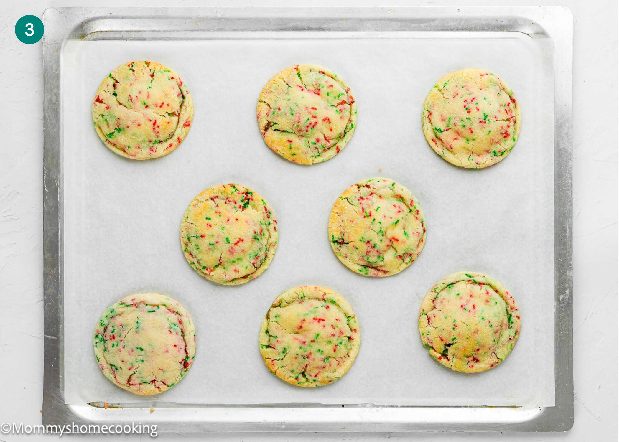 A baking tray with eight round Eggless Christmas Sprinkle Sugar Cookies topped with colorful sprinkles on parchment paper, fresh out of the oven.