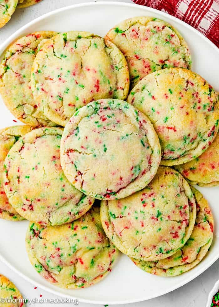 A plate of Eggless Christmas Sprinkle Sugar Cookies with red and green sprinkles, arranged in a pile on a white dish.