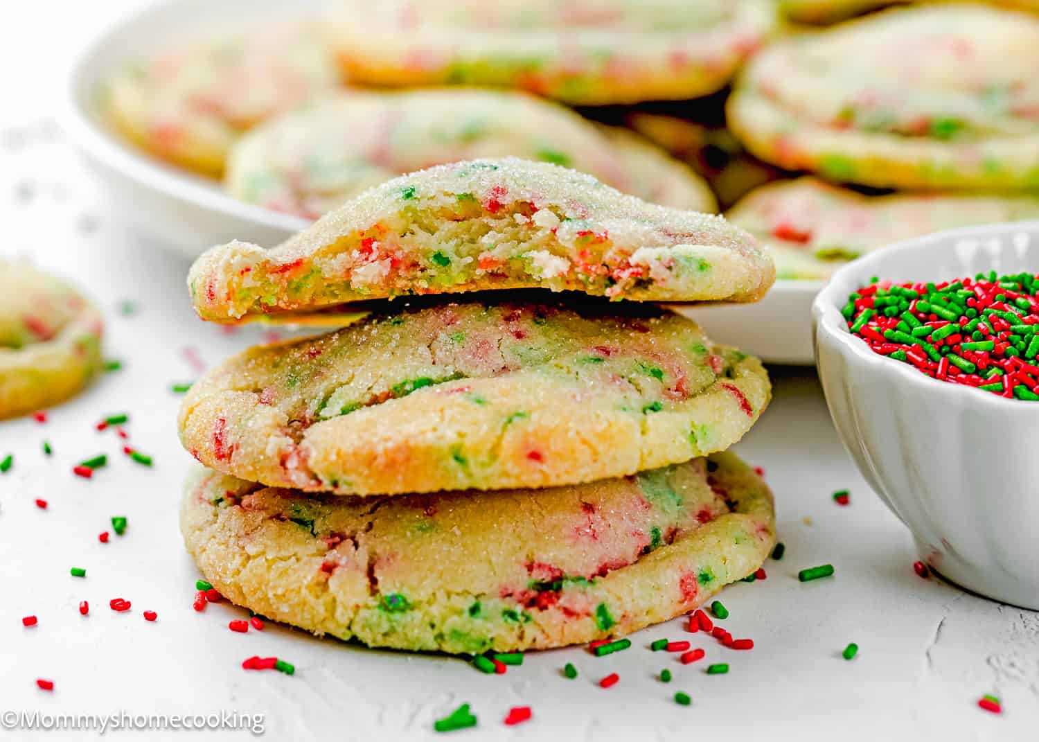 Three Eggless Christmas Sprinkle Sugar Cookies with red and green sprinkles are stacked on a white surface, with more cookies and a bowl of sprinkles in the background.