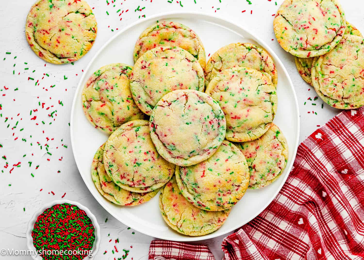 A white plate filled with Eggless Christmas Sprinkle Sugar Cookies with red and green sprinkles, surrounded by a small bowl of sprinkles, cookies, and a red plaid cloth on a white surface.