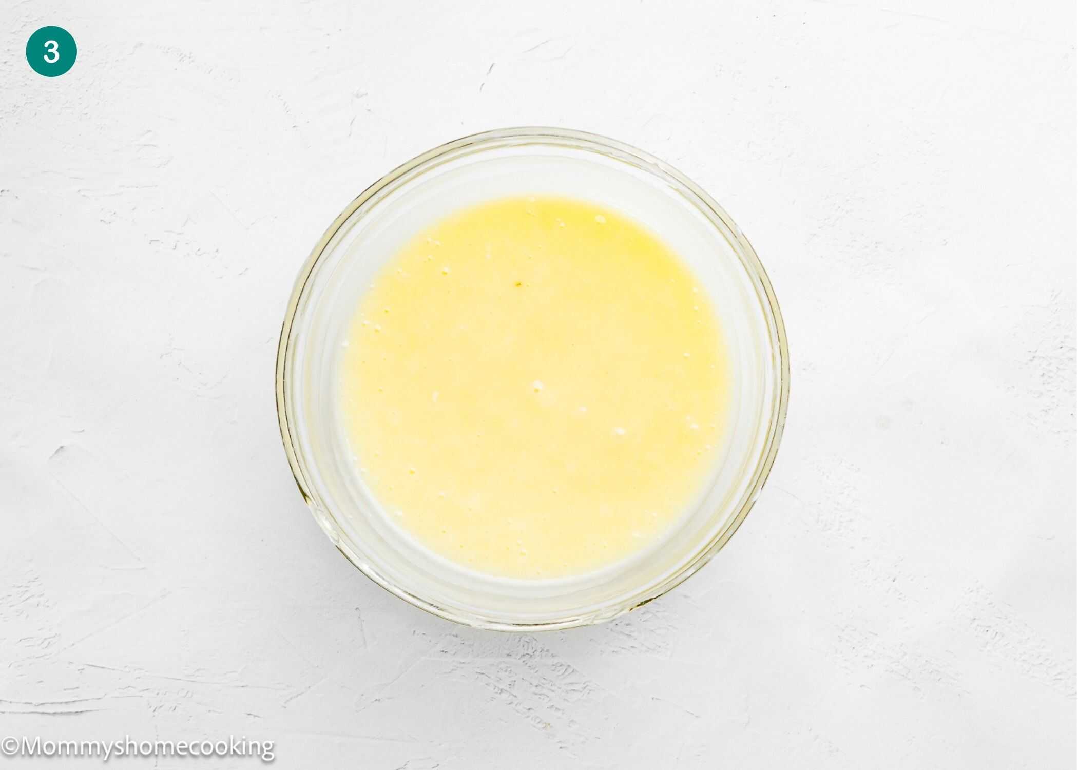 A clear glass bowl containing a yellow liquid mixture for Eggless Cinnamon Roll Pancakes is placed on a white surface. The number 3 is shown in a blue circle in the top left corner.