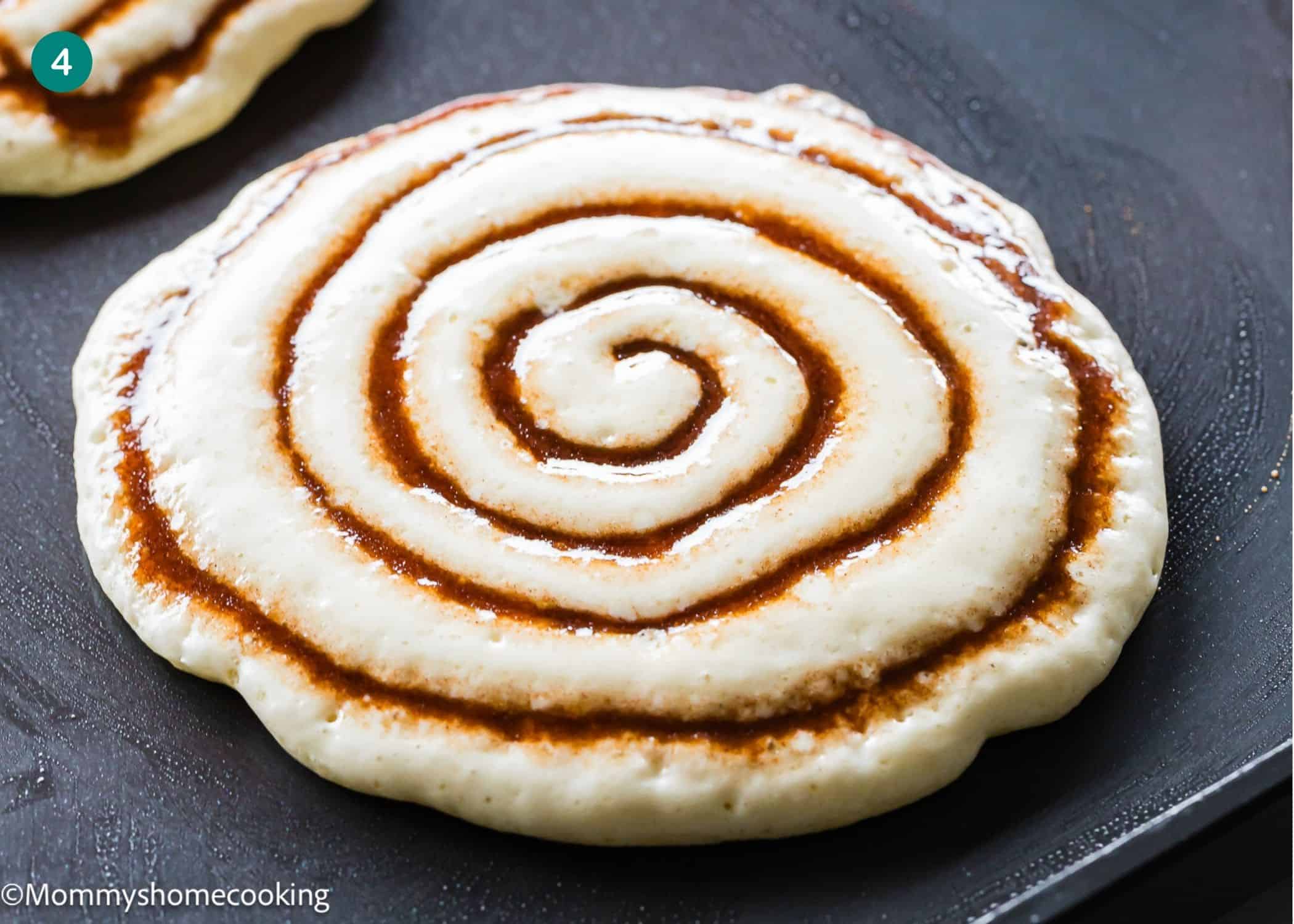 An Eggless Cinnamon Roll Pancake cooks on a griddle, its surface adorned with a spiral swirl of cinnamon filling.