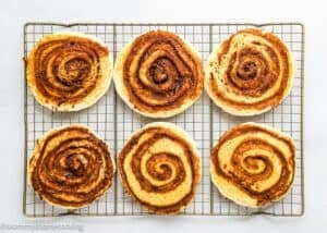 Six cinnamon rolls arranged in two rows on a wire cooling rack over a white surface, perfect for pairing with Eggless Cinnamon Roll Pancakes 14 for a delightful breakfast spread.