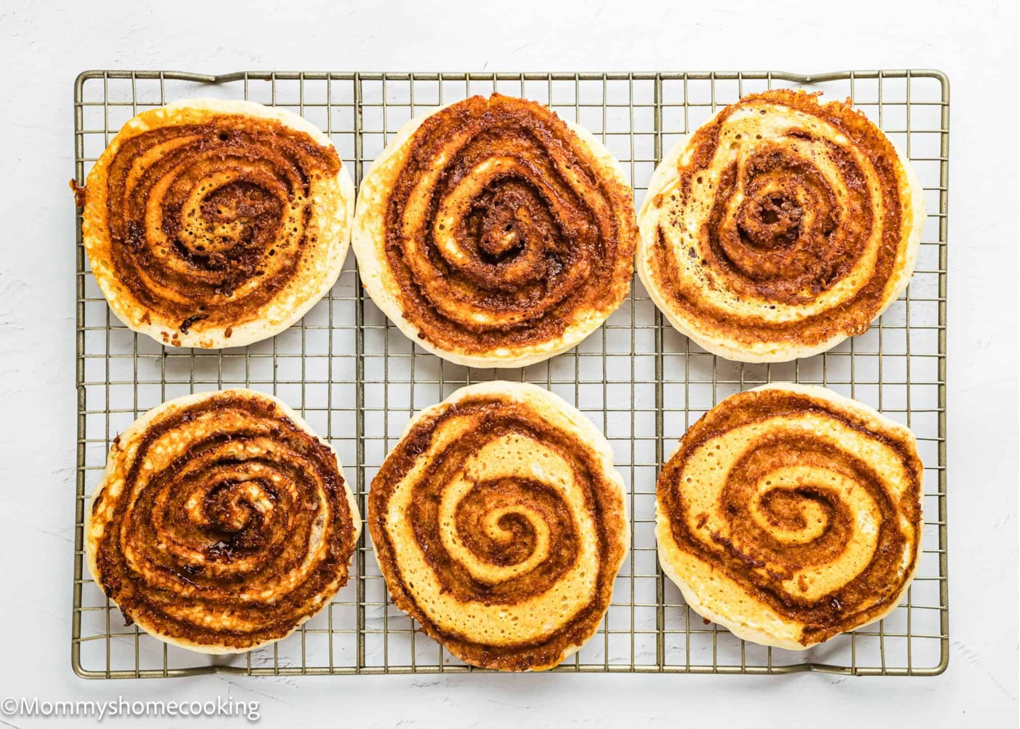 Six cinnamon rolls arranged in two rows on a wire cooling rack over a white surface, perfect for pairing with Eggless Cinnamon Roll Pancakes 14 for a delightful breakfast spread.