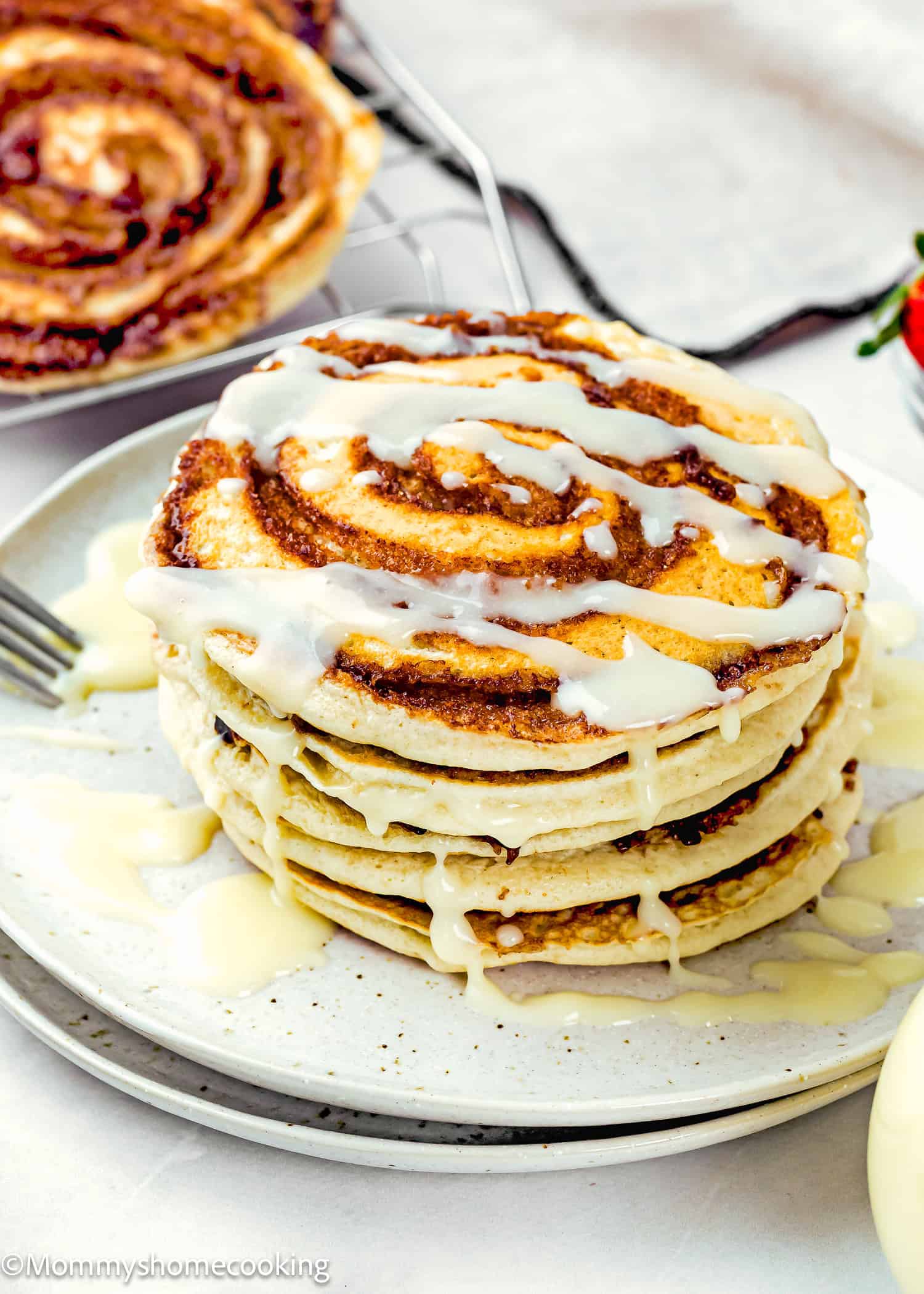 A stack of Eggless Cinnamon Roll Pancakes drizzled with white icing sits on a plate, with a fork and more pancakes in the background.
