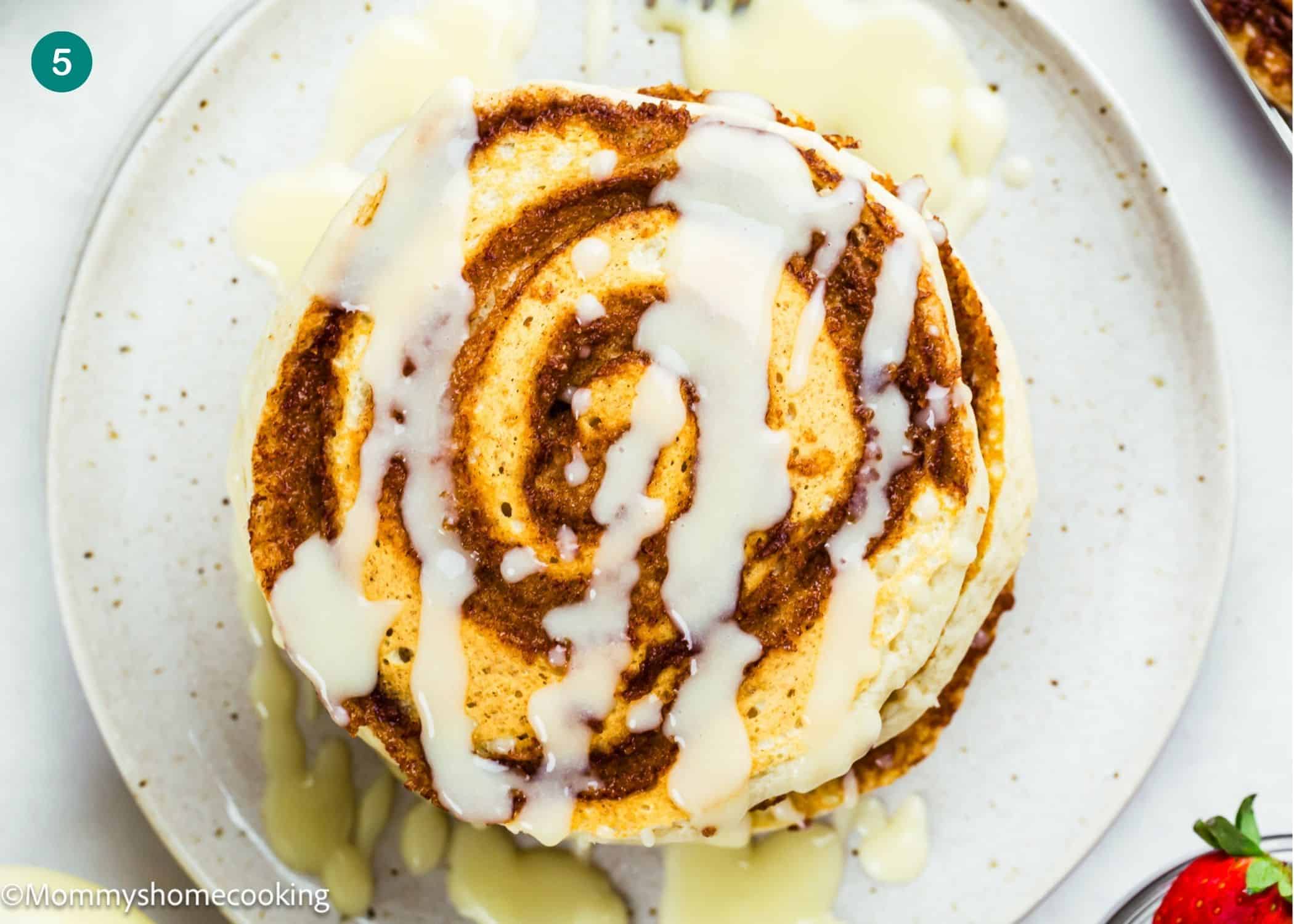 A stack of Eggless Cinnamon Roll Pancakes topped with white icing, served on a white plate.