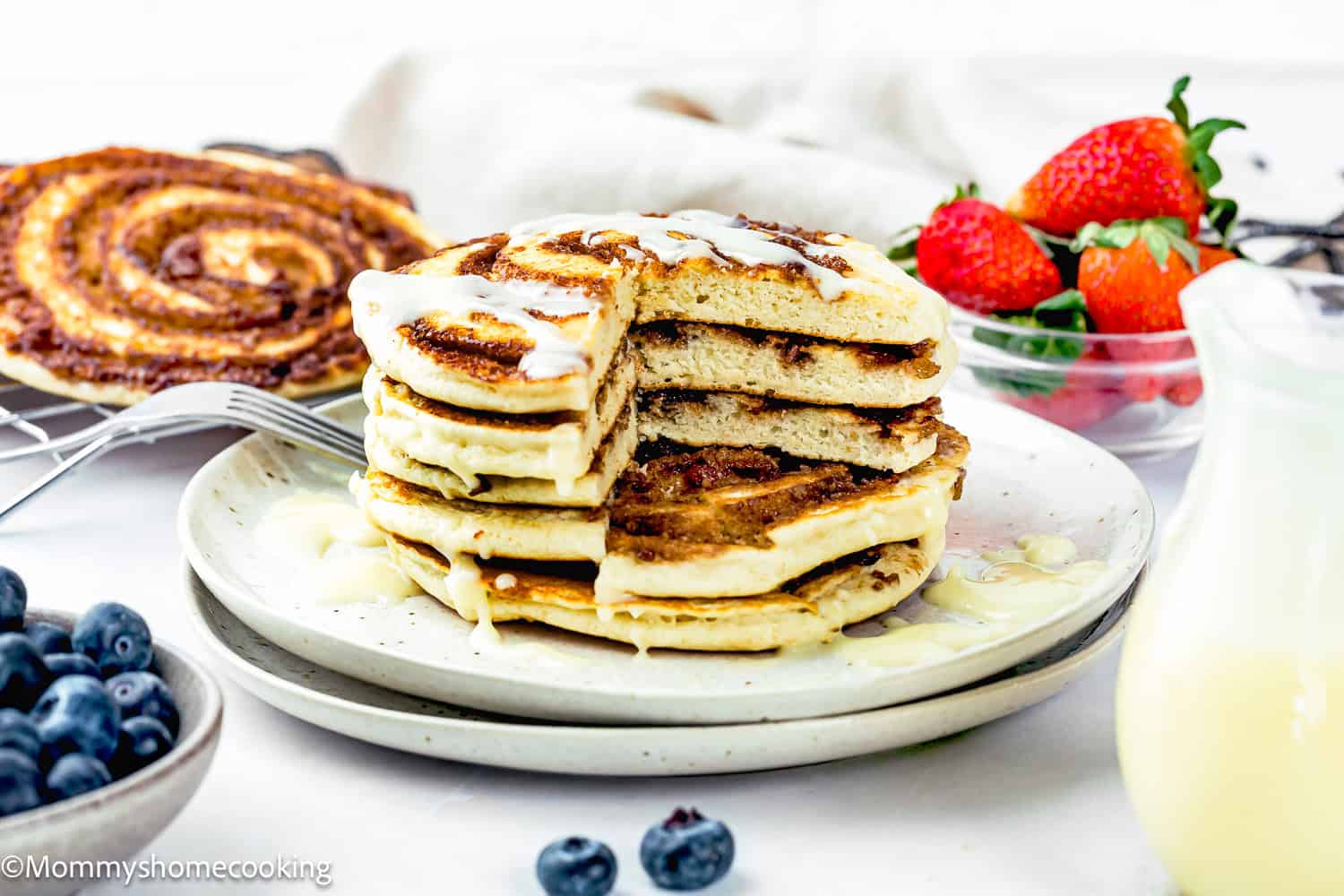 A stack of Eggless Cinnamon Roll Pancakes drizzled with icing, served on a plate with fresh blueberries and strawberries.