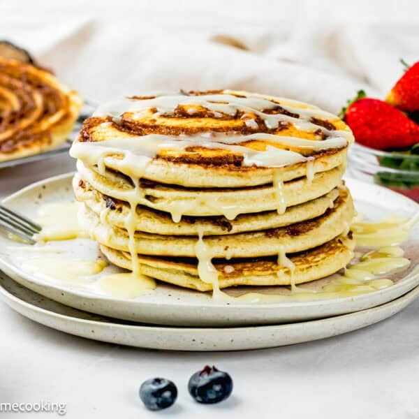 A stack of Eggless Cinnamon Roll Pancakes drizzled with icing sits on a plate, with a bowl of strawberries and some blueberries beside it.