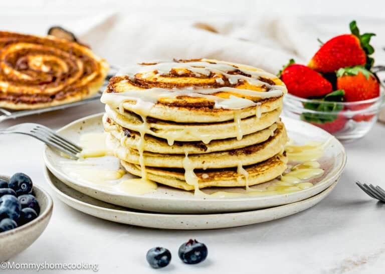 A stack of Eggless Cinnamon Roll Pancakes drizzled with icing sits on a plate, with a bowl of strawberries and some blueberries beside it.