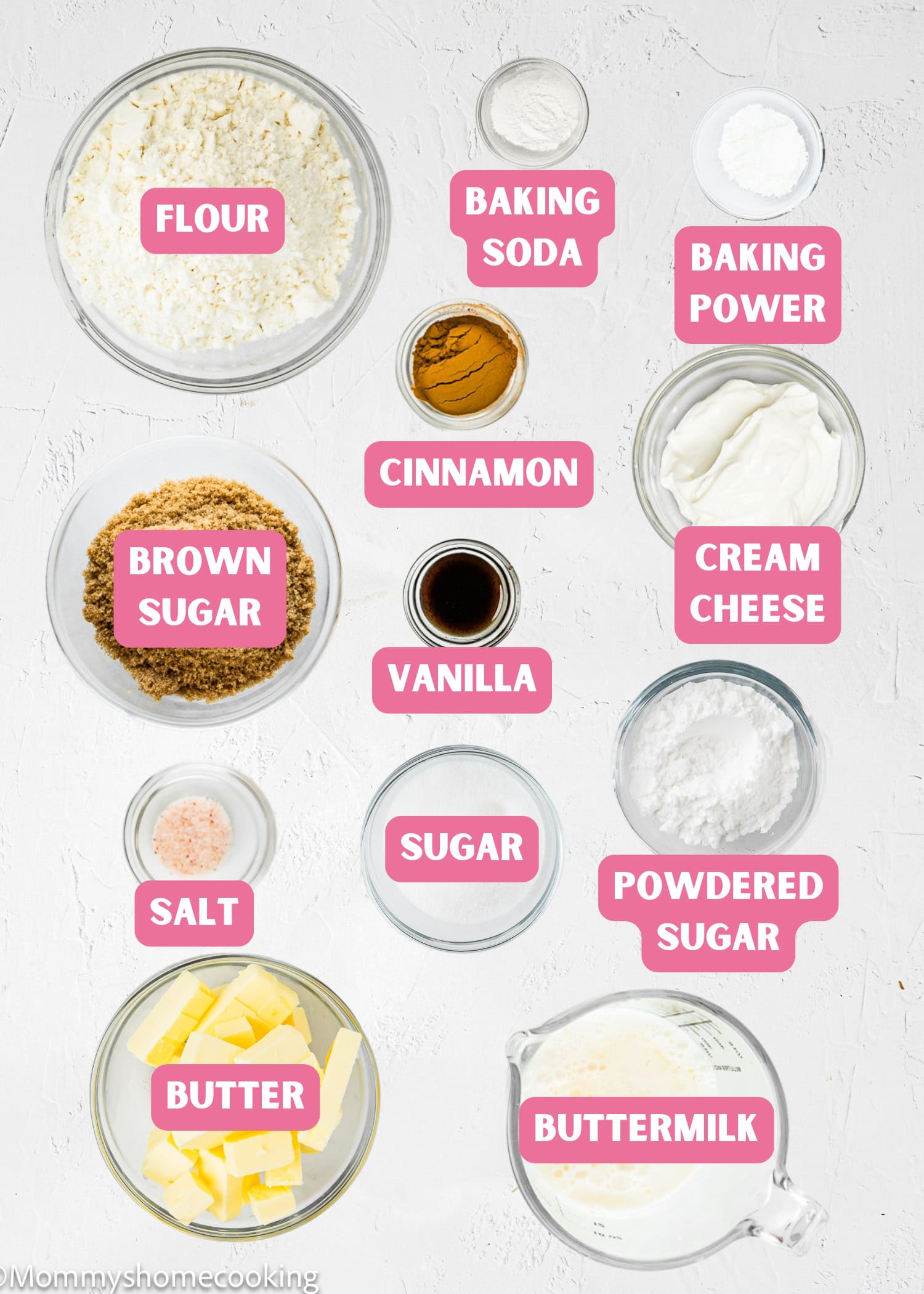 Top-down view of labeled baking ingredients in bowls for Eggless Cinnamon Roll Pancakes, including flour, brown sugar, baking soda, baking powder, cinnamon, cream cheese, vanilla, sugar, powdered sugar, butter, salt, and buttermilk.