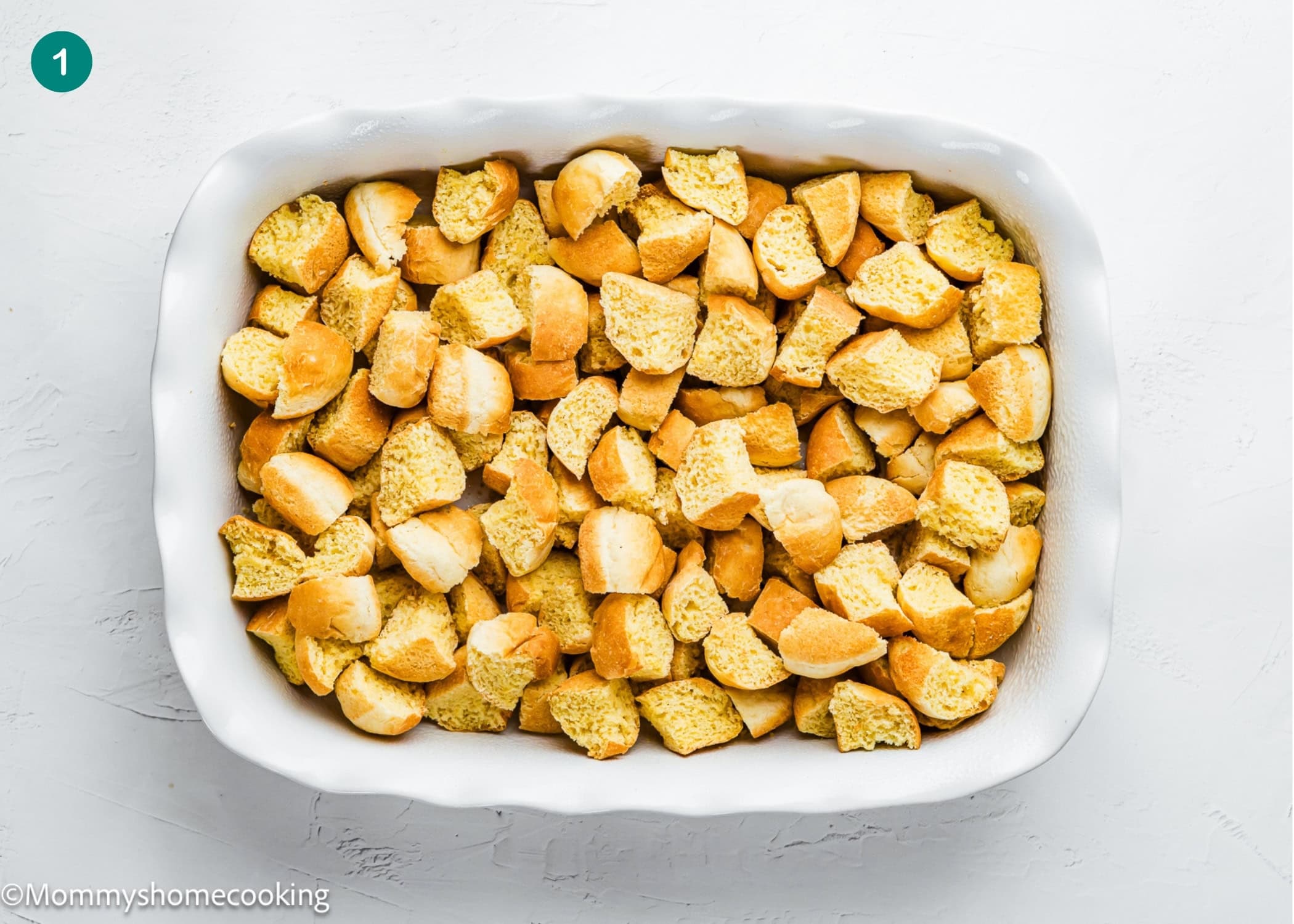A white baking dish filled with evenly spread chunks of cubed bread on a white surface, perfect for preparing Overnight French Toast Casserole Without Eggs.