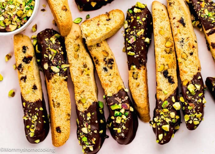 Rows of biscotti from an Eggless Easy Biscotti Recipe are partially dipped in chocolate and sprinkled with chopped pistachios, arranged on a light surface with a bowl of extra pistachios nearby.
