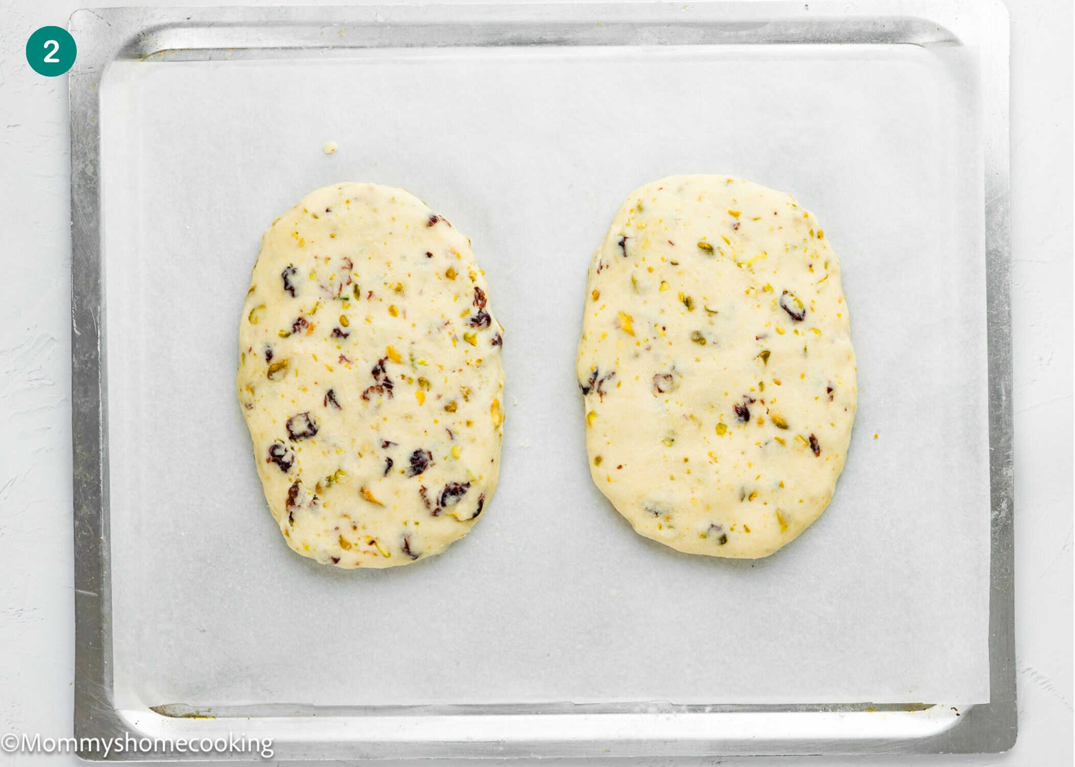 Two oval-shaped, unbaked biscotti doughs with visible cranberries and nuts are placed on a parchment-lined baking sheet, ready for this Eggless Easy Biscotti Recipe.