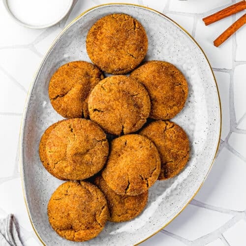 A white oval plate with nine Eggless Gingerbread Snickerdoodle cookies, surrounded by jars of sugar and cinnamon powder, and two cinnamon sticks on a white tiled surface.