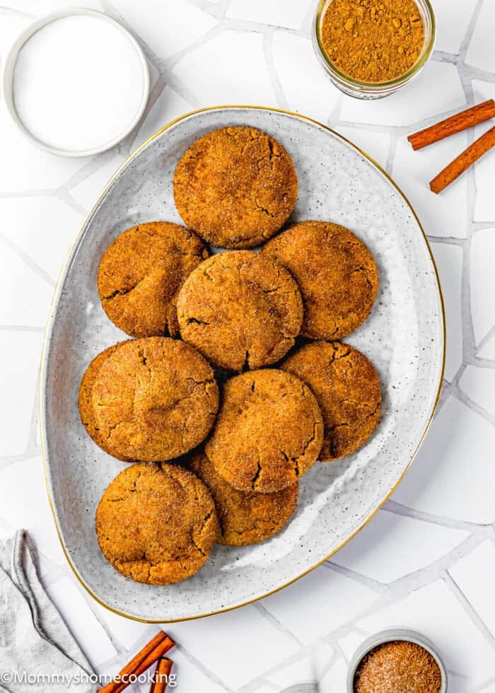 A white oval plate with nine Eggless Gingerbread Snickerdoodle cookies, surrounded by jars of sugar and cinnamon powder, and two cinnamon sticks on a white tiled surface.