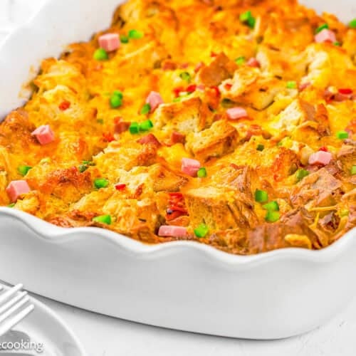 An Eggless Strata Recipe baked in a white dish, topped with diced ham, green onions, and melted cheese.