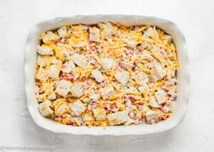 A rectangular baking dish filled with a mixture of cubed bread, shredded cheese, diced ham, and chopped vegetables showcases this tasty Eggless Strata Recipe on a white surface.