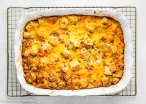 A baked Eggless Strata Recipe breakfast casserole with bread cubes, diced ham, cheese, and green peppers in a white rectangular dish on a cooling rack.
