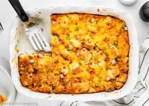 An Eggless Strata Recipe baked in a white dish with melted cheese, bread cubes, diced ham, and green onions, featuring a metal spatula and one serving removed.