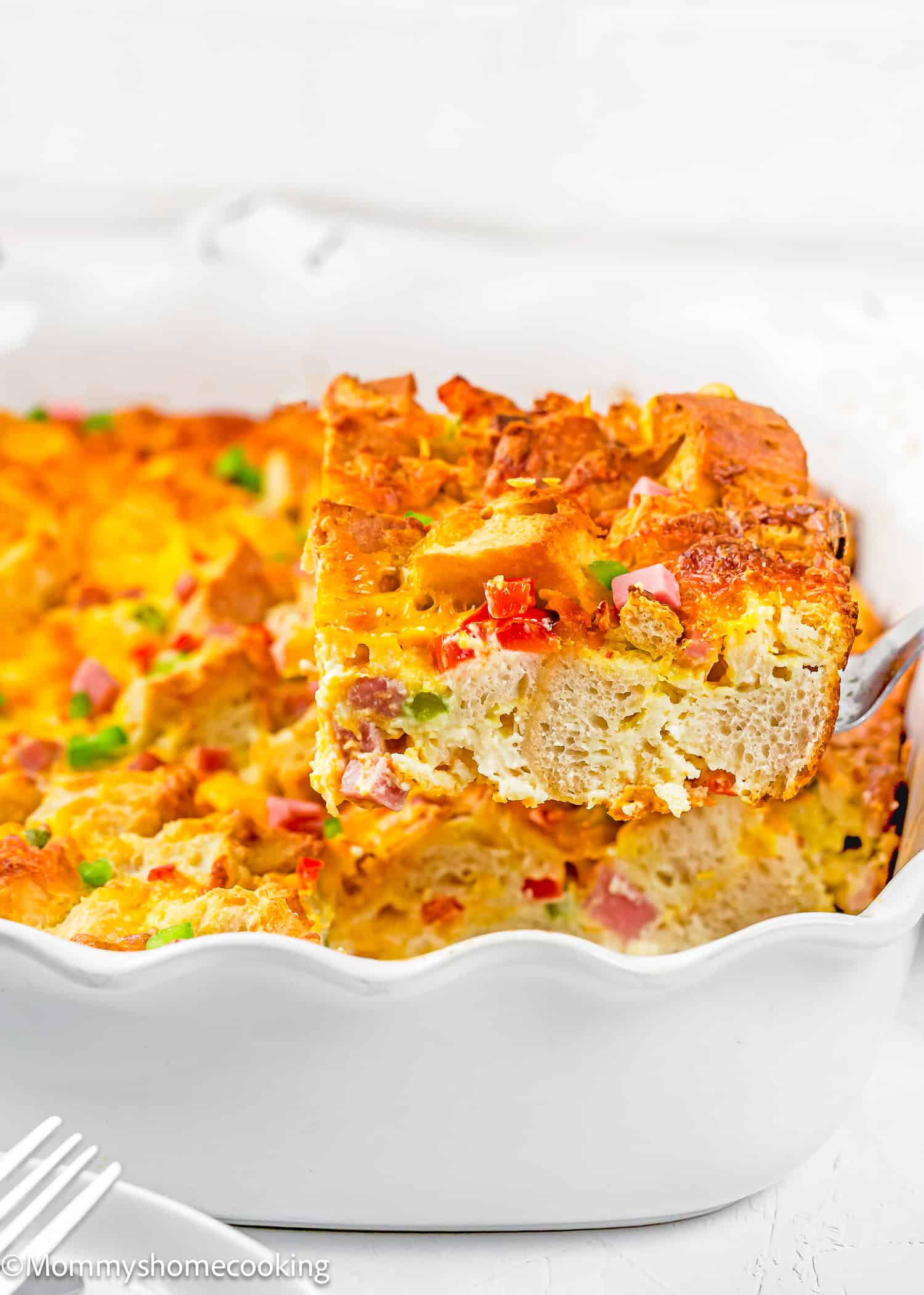 A fork lifts a square slice of Eggless Strata Recipe with bread, ham, cheese, and vegetables from a white baking dish.