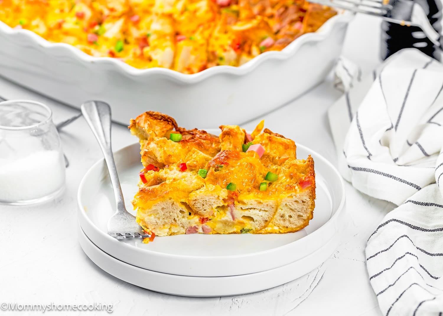 A slice of breakfast casserole made with bread, cheese, and vegetables on a white plate with a fork; try this Eggless Strata Recipe for a delicious start to your day. Casserole dish and napkin in the background.