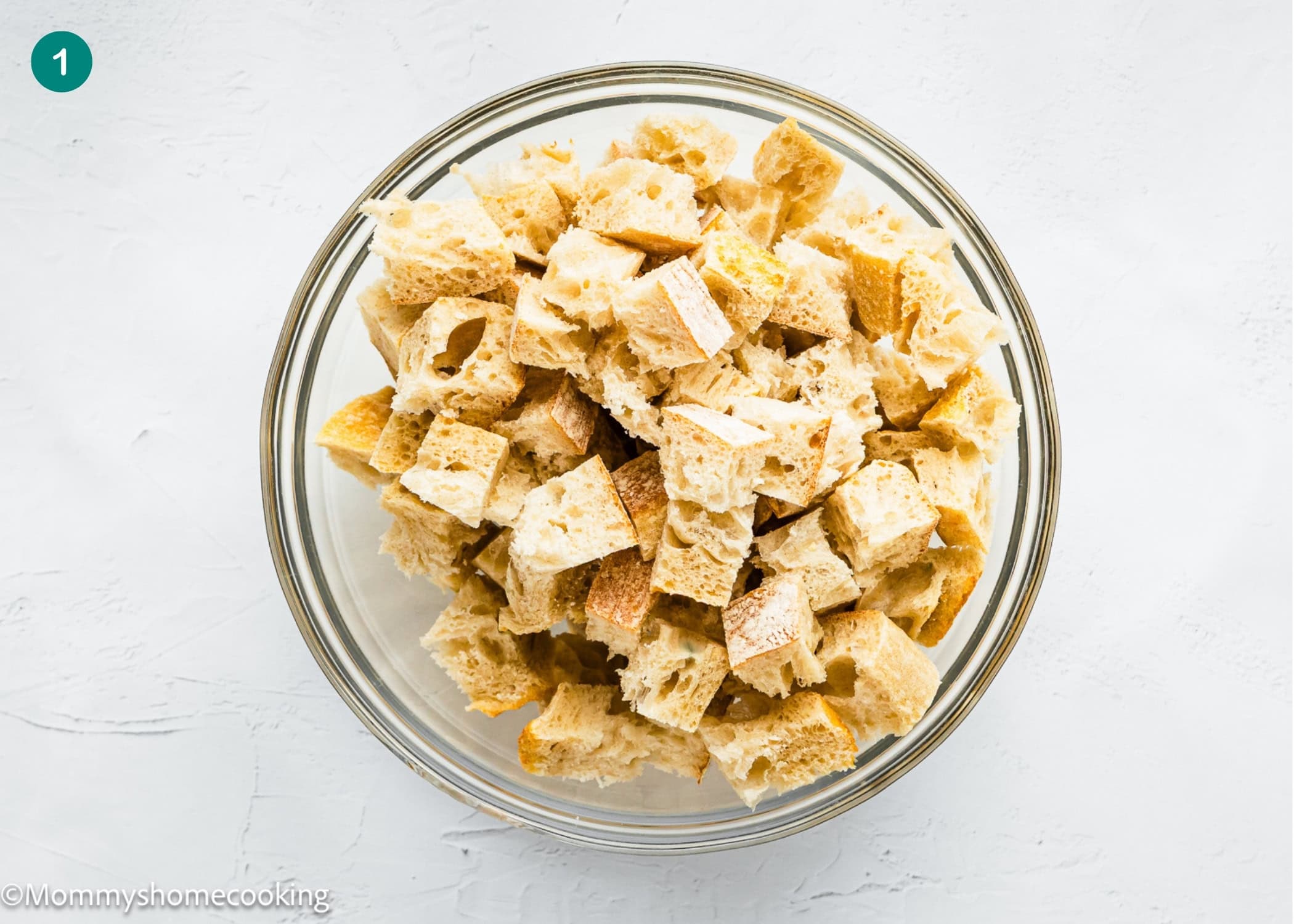 A glass bowl filled with cubed pieces of bread on a white surface, perfect for preparing an Eggless Strata Recipe.