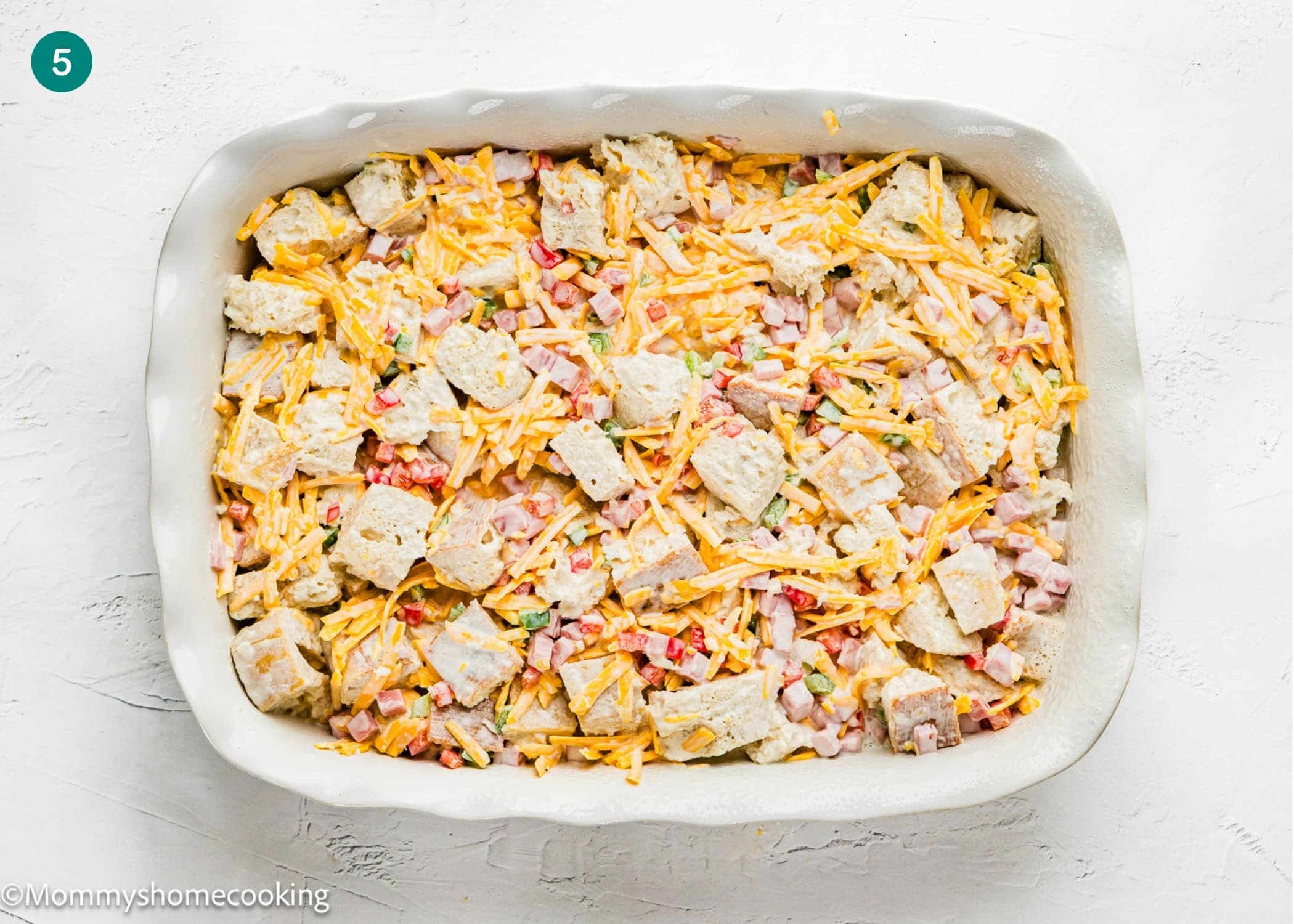 A white baking dish filled with a mixture of cubed bread, shredded cheese, diced ham, and chopped vegetables sits on a white surface—perfect for an easy Eggless Strata Recipe.