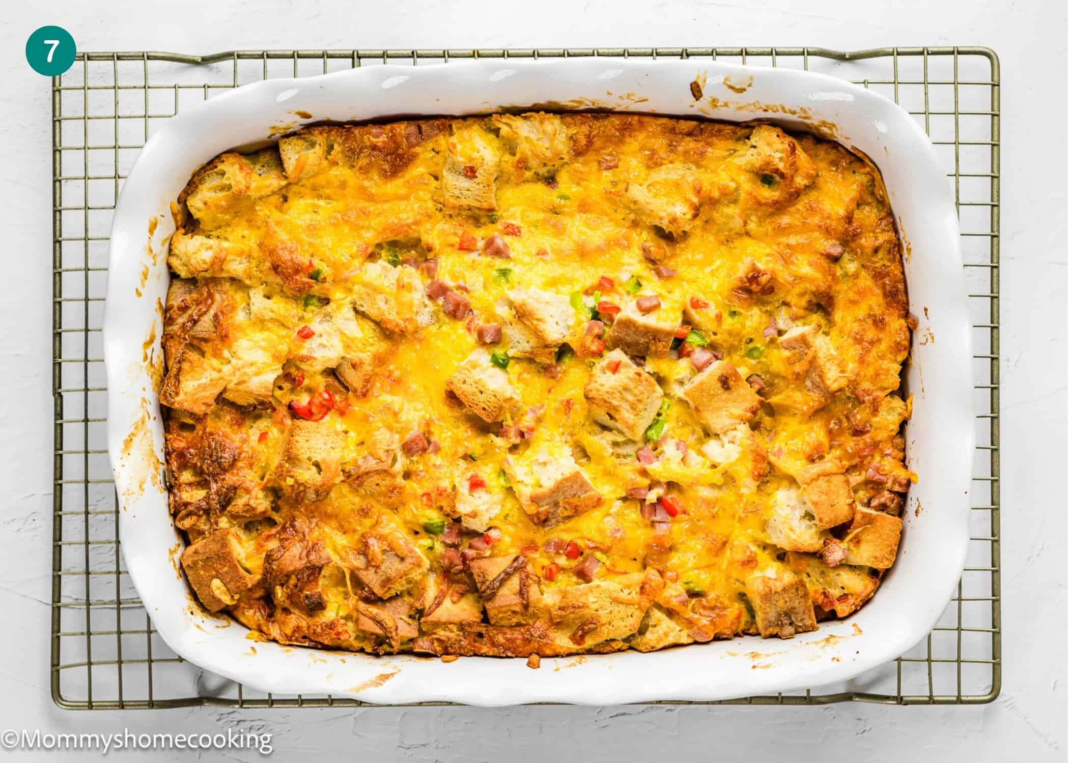 An Eggless Strata Recipe featuring cubed bread, melted cheese, and chopped vegetables baked in a white rectangular dish on a cooling rack.