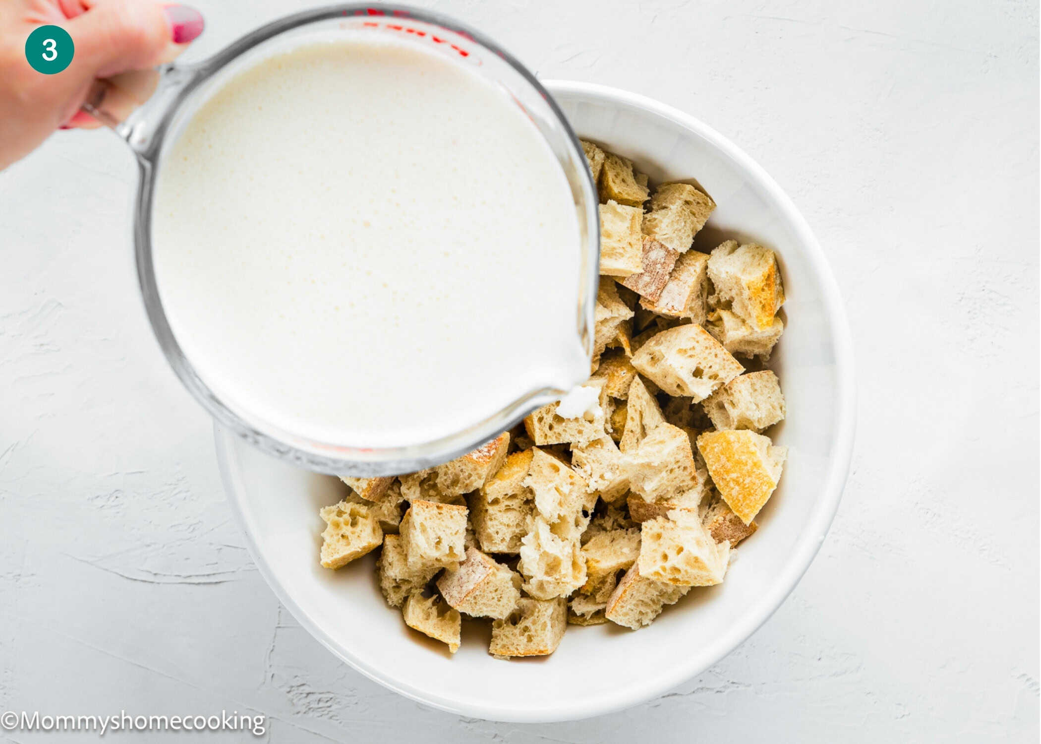 A measuring cup of milk mixture is being poured over a bowl of cubed bread pieces on a white surface, perfect for an Eggless Strata Recipe.